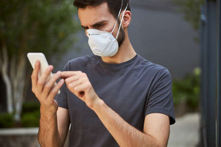 Portrait of a serious young male in a protective mask writing a text message on his cellphone
