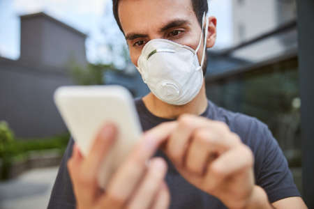 Close up portrait of a focused guy in a disposable face mask reading a text messageの写真素材
