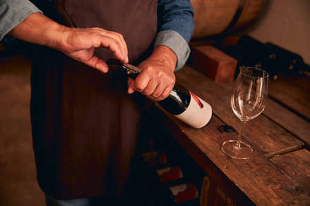 Close up of male hands using corkscrew to open bottle of alcoholic drink while man standing by wooden table with empty wineglassの写真素材