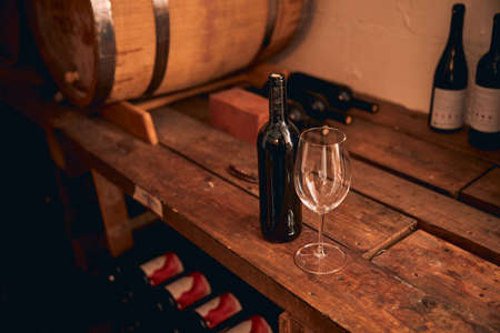 Wineglass and bottle of alcoholic drink on wooden shelf with barrel in wine cellarの写真素材