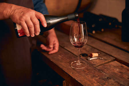 Close up of male hand pouring alcoholic drink into wineglass while man standing by wooden table in...