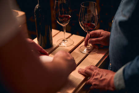 Cropped photo of unrecognized men standing near the wooden crate with glasses of red wine on it