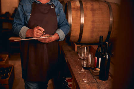 Cropped photo of a man in a brown apron standing with a clipboard and a pen next to a wine barrel. Bottles and glasses on the tableの写真素材