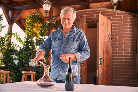 Cheerful man in a denim shirt standing on a terrace and smiling while pouring red wine into a glass...