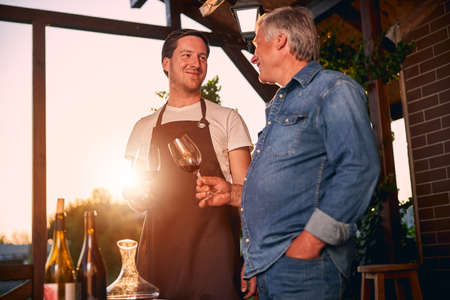Cheerful young sommelier in a brown apron clanging glasses with an older colleague on a winery...