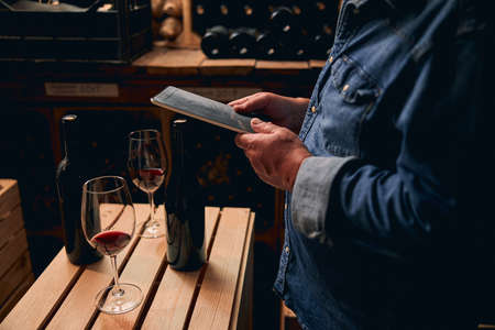 Cropped photo of a man in a denim shirt standing with a tablet in winery cellar. Glasses and bottles...