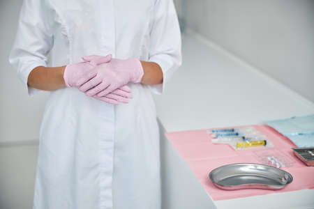 Cropped photo of a Caucasian female cosmetologist in latex gloves standing by an instrument tableの写真素材
