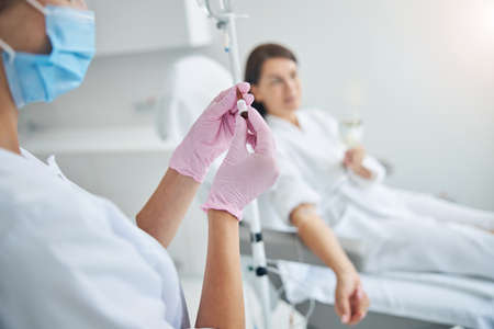 Nurse in a face mask standing by a dark-haired female patient during the intravenous therapy