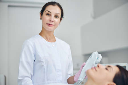 Smiling pleased certified middle-aged cosmetologist standing by a Caucasian female patient during a cosmetic procedureの写真素材