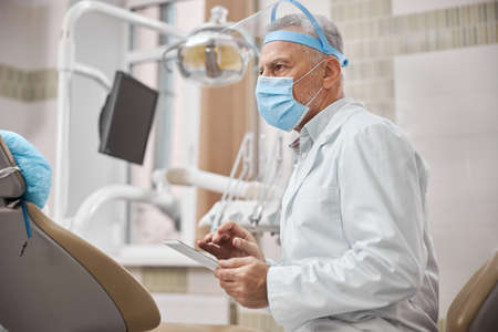 Experienced elderly dentist working on his tablet while waiting for a patient in his workplaceの写真素材