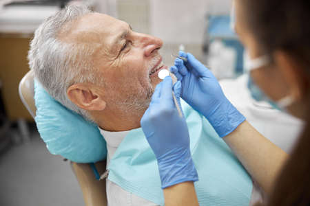 Professional dental specialsit holding a mouth mirror and a dental pick while examining an elderly...