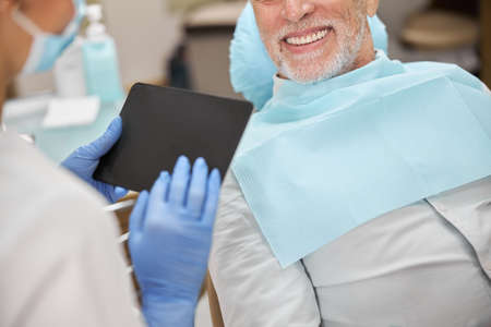 Cropped photo of a dental expert holding a tablet while having an appointment with an elderly man
