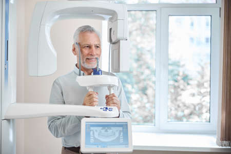 Grey-haired man smiling while standing by a panoramic radiographer in a dental clinic