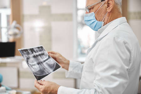 Professional aging dental expert wearing a surgical mask and looking at a dental x-ray image