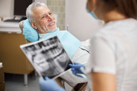 Joyful elderly man sitting in a dental chair looking at a professional female dentist holding an x-ray imageの写真素材