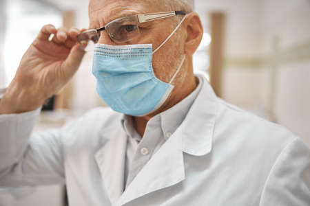 Close-up photo of a professional elderly healthcare worker touching his glasses and looking serious at workの写真素材
