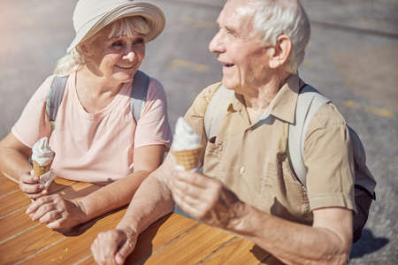Joyful aged lady and her husband sitting at a wooden table with ice cream cones in their handsの写真素材