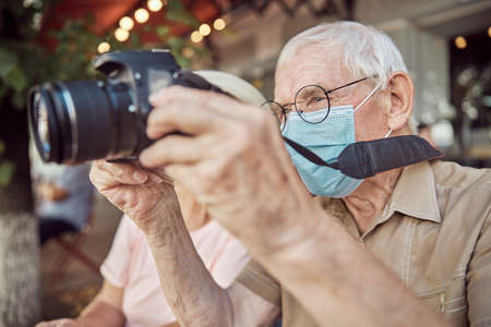 Focused aged Caucasian man in eyeglasses and a face mask photographing with his digital cameraの写真素材