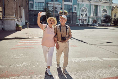 Merry aged woman photographing herself and her husband with a smartphone at the pedestrian crossingの写真素材