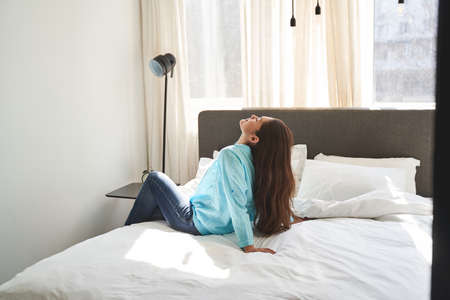 Side view of a dark-haired lady with her head thrown back sitting on the bedの写真素材