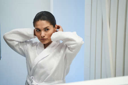 Waist-up portrait of a focused woman in a bathrobe staring at herself in the mirror