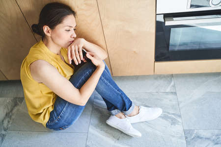 Side view of a joyless young woman sitting on the tile floor in the kitchen