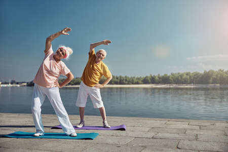 Full-length portrait of a smiling senior lady and her spouse doing a side bend exercise