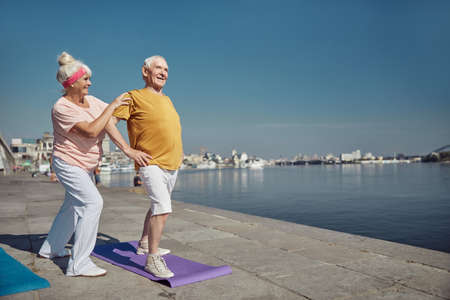 Pleased male pensioner doing a standing shoulder stretch assisted by a joyous senior Caucasian ladyの写真素材