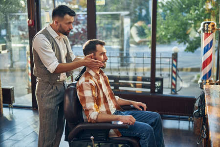 Attentive barber standing behind his client and holding his head while looking in the mirror