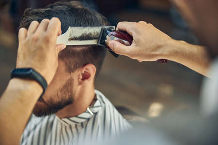 Close-up photo of electric hair clipper and a hair comb in hands of a skillful barber