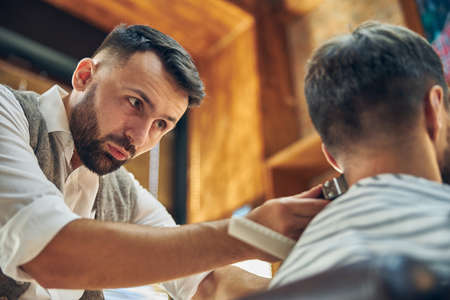 Concentrated hair-stylist trimming hair of his visitor with electric clippers at a barbershopの写真素材