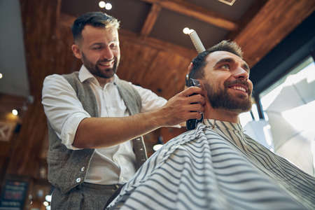 Ambitious hair-stylist smiling while trimming hair of his joyful young client