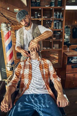 Serious barber holding a straight razor to the neck of his client while shaving him and and looking at the cameraの写真素材