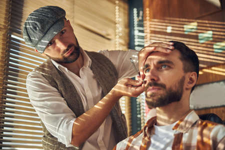 Professional young hairdresser wearing a cool hat and shaving his client while looking at the cameraの写真素材
