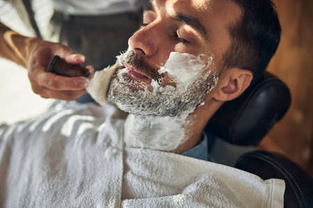 Close-up photo of a brunette man keeping eyes closed while having shaving foam put on his face at a barbershopの写真素材