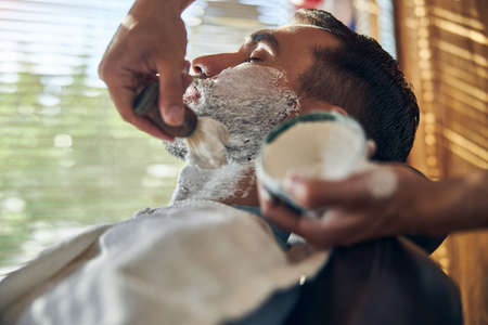 Calm man with eyes closed being covered in shaving cream during an appointment at a barbershopの写真素材