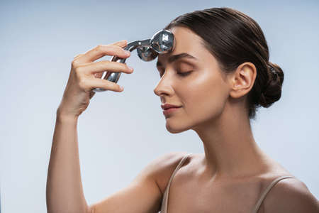 Side view portrait of beautiful brunette woman using metal roller on forehead isolated on light blue...