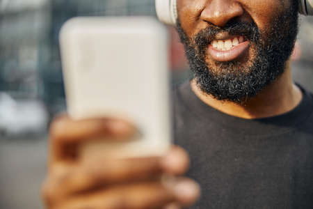 Cropped photo of a bearded man holding a modern smartphone and revealing his teeth while smiling