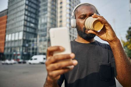 Calm young man drinking coffee from a paper cup in the street and looking at the smartphone screenの写真素材