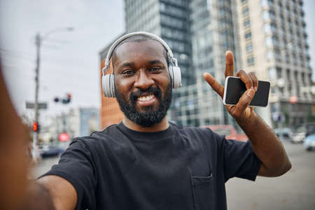 Cheerful bearded young man wearing headphones holding a modern smartphone and showing peace gesture with a happy smileの写真素材