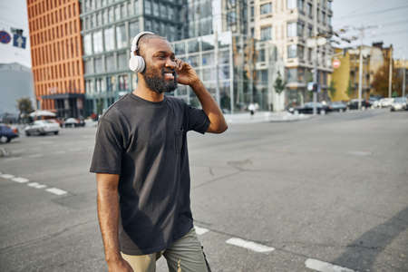 Contented young man in casual clothes walking in the city center and touching the wireless...