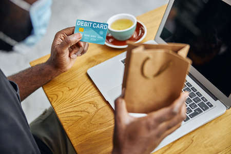 Cropped photo of a debit card and a paper gift bag in hands of Afro American man sitting at the...