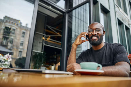 Handsome bearded man wearing glasses smiling while sitting at the outdoors cafe table and enjoying a pleasant phone talk. Website bannerの写真素材