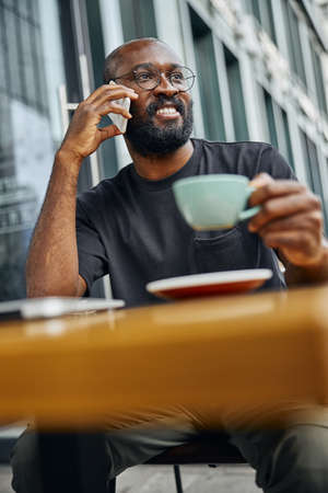 Relaxed joyful man sitting at the outdoor table with a cup and smiling while talking on the phoneの写真素材