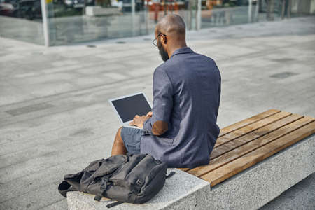 Serious male person sitting on the bench and using his laptop for typing answer on the emailの写真素材