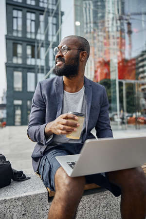 Handsome bearded man keeping smile on his face while sitting with laptop on the benchの写真素材