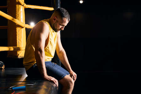 Thoughtful fighter dangling his legs from the edge of the boxing ring and looking downの写真素材