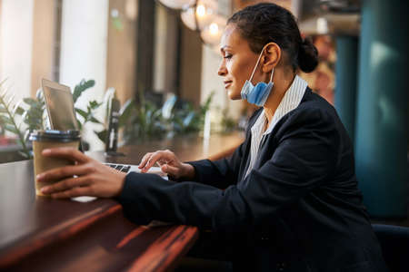 Pleased dark-skinned woman using her laptop while working on distance from cafeの写真素材