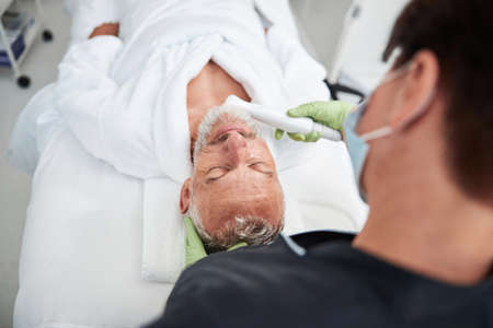 Competent female cosmetologist doing a skin rejuvenation procedure for an aged man with a pulsed light apparatusの写真素材