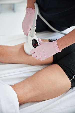 Professional cosmetologist wearing gloves and pressing an ultrasonic machine to legs of a pantless...